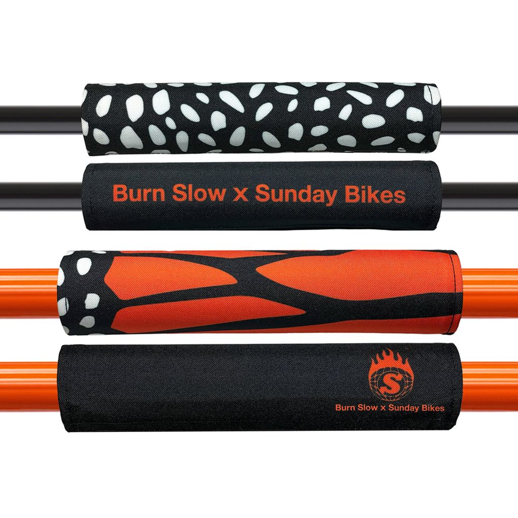 The Sunday X Burn Slow Reversible Handlebar & Frame Pad set includes four handlebar pads and a reversible frame pad featuring black and white patterns, plus an orange design with "Burn Slow x Sunday Bikes" branding, enhancing style and offering essential knee protection.