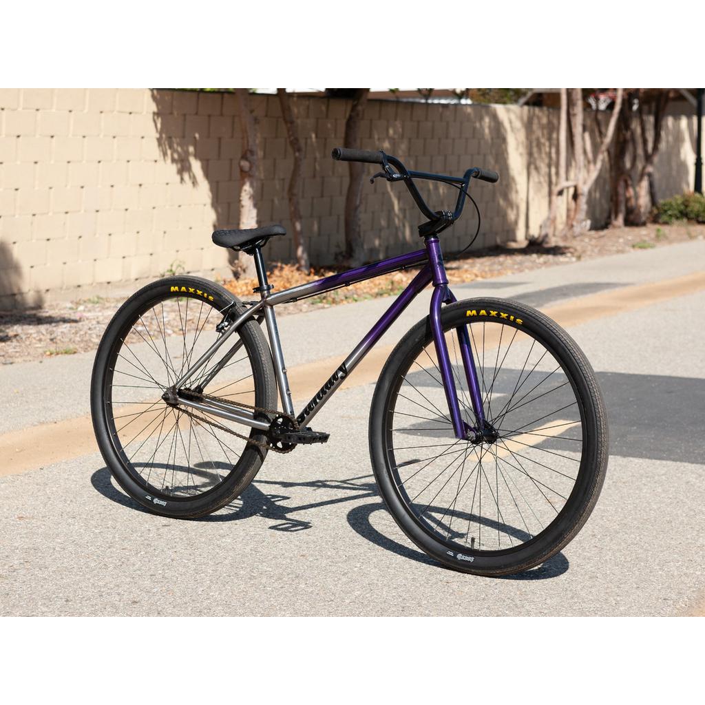 The Sunday High C 29 Inch Bike, a purple and silver bicycle with black handlebars, Maxxis tires, and a chromoly frame, is parked on a paved road near a beige brick wall.