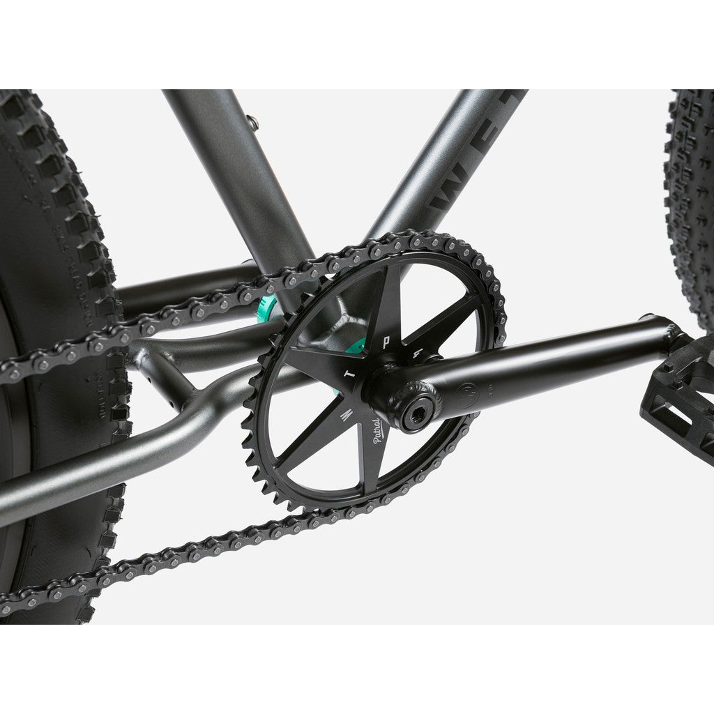 Close-up of the chainring, crank, and chain on the sleek black frame of the Wethepeople Avenger 27.5 Inch Bike. The rear wheel's textured tire suggests its hybrid clunker machine design with powerful disc brakes for superior control.