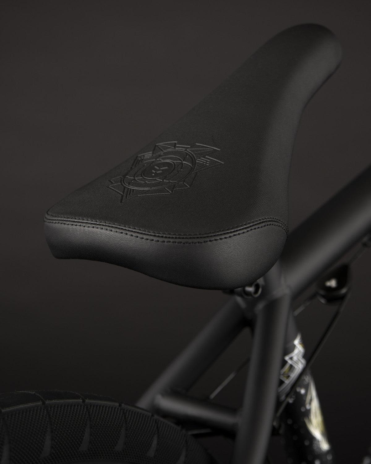 A close-up of the Fly Bikes Electron 20 Inch Bike features a black bicycle seat with an embossed design, mounted on a Cr-Mo frame, set against a dark background.