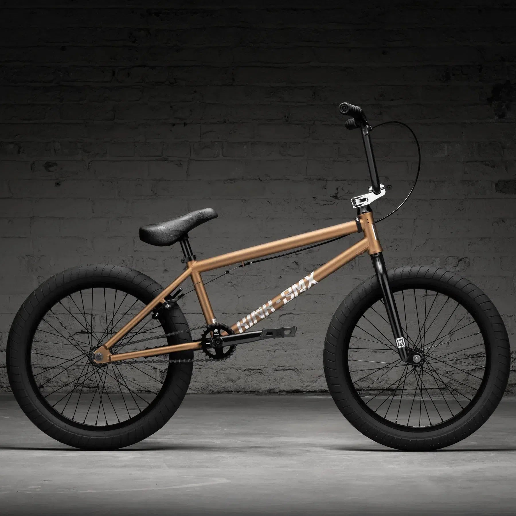 A tan Kink Launch 20 Inch Bike (2026) with thick black tires and a sleek black seat stands against a dark brick wall, showcasing its precision with high-quality sealed bearings.