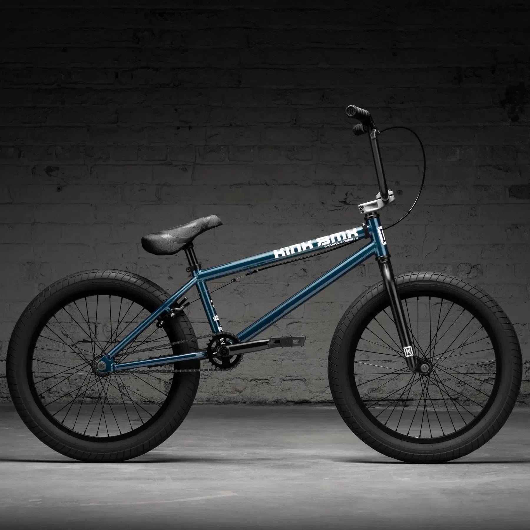 The Kink Curb 20 Inch Bike (2026), a sleek blue BMX with wide black tires, is parked indoors on a concrete floor against a gray brick wall, ideal for entry-level riders.