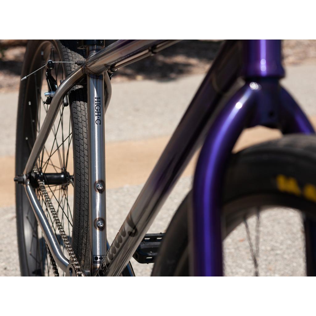 A close-up of the Sunday High C 29 Inch Bike highlights its rear wheel and gleaming chromoly frame shining brightly in the sunlight against a blurred background.