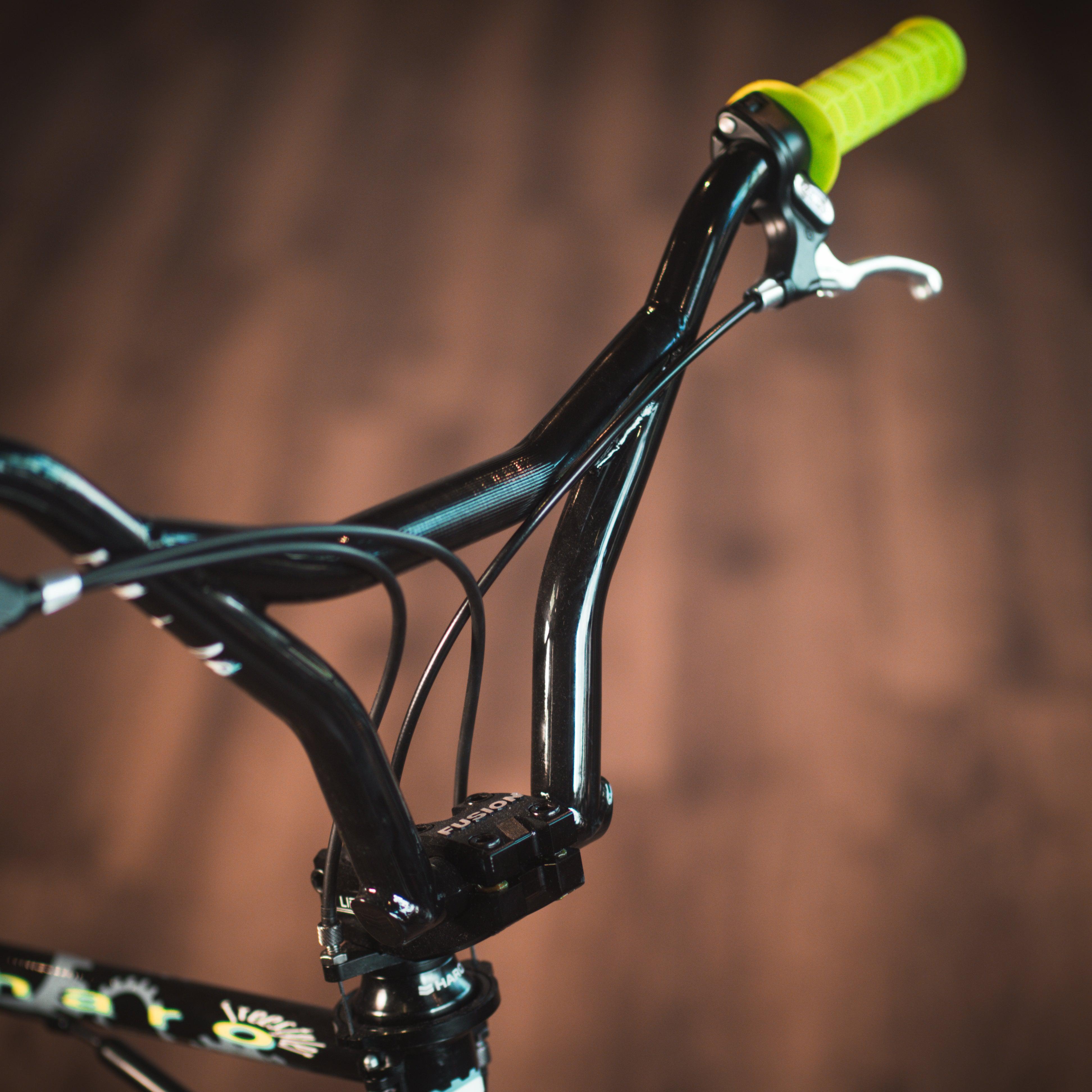 Close-up of the handlebar of the Haro Lineage Sport Freestyler Bashguard 26 Inch Bike, featuring yellow grips and a brake lever, with a brown wooden floor in the background.