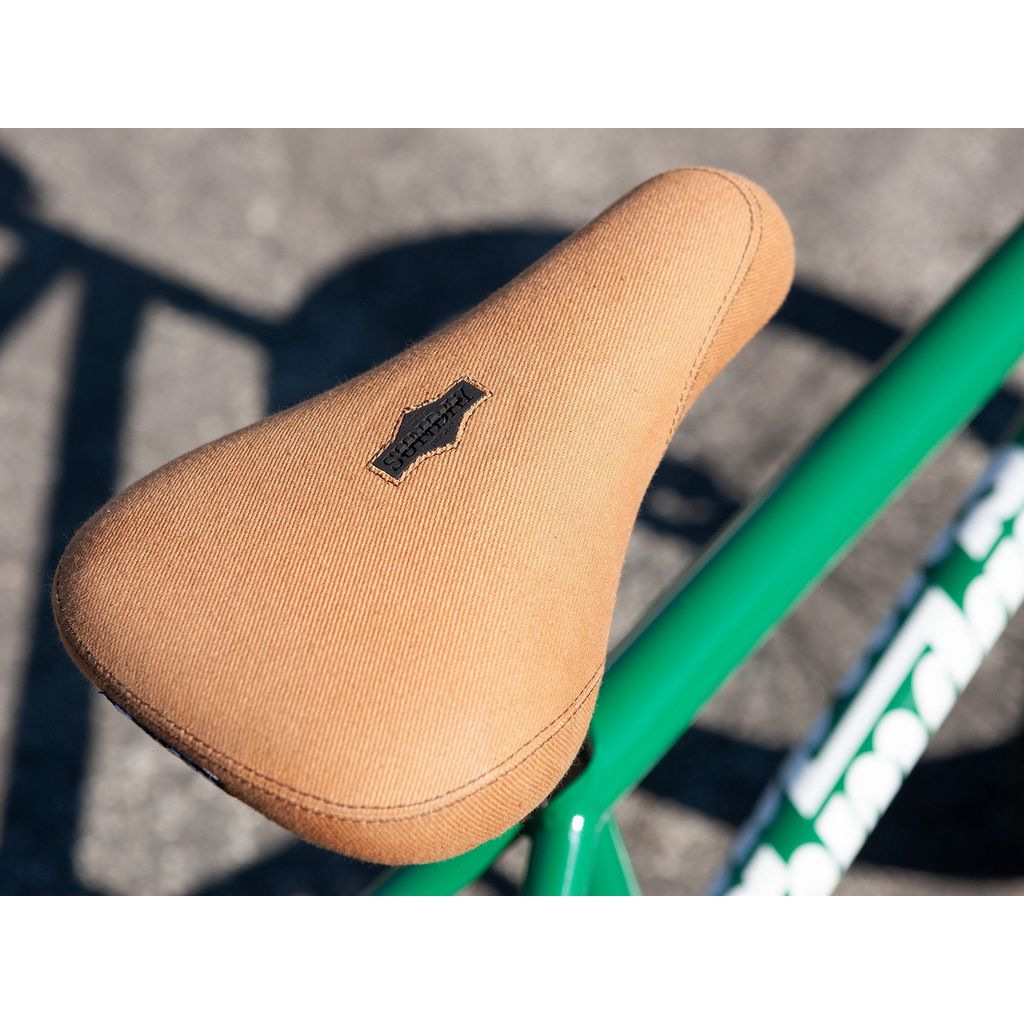 Close-up of a bicycle saddle with a brown fabric cover, mounted on a green Sunday Forecaster 20 Inch Bike (2022) / Alec Siemon frame, casting a shadow on the pavement in the background.