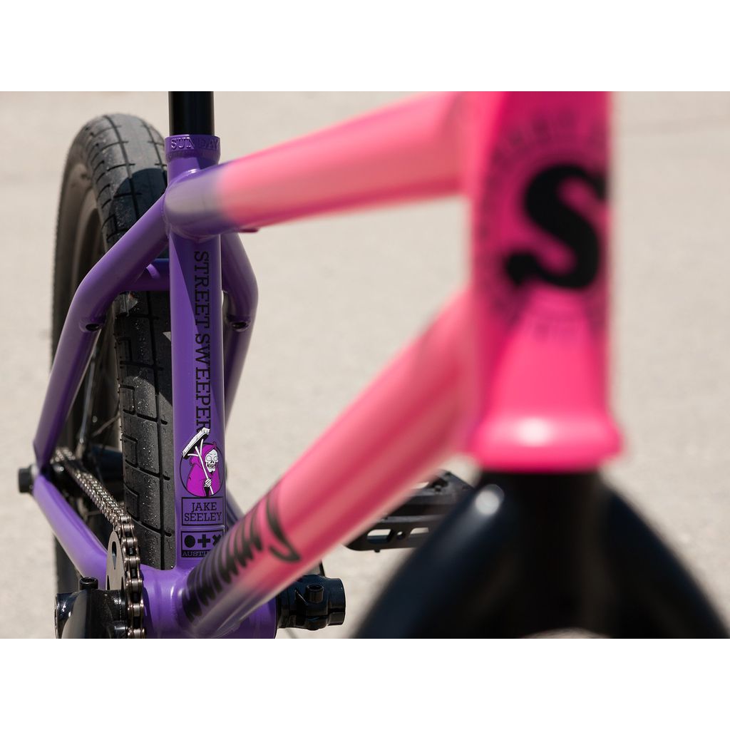 A detailed view of the Sunday Street Sweeper 20 Inch Bike frame, expertly constructed from durable chromoly, showcases a bright blend of purple and pink shades. Featuring a "Street Sweeper" decal alongside a whimsical purple cartoon character at its center, this Jake Seeley signature model is designed to stand out with flair.