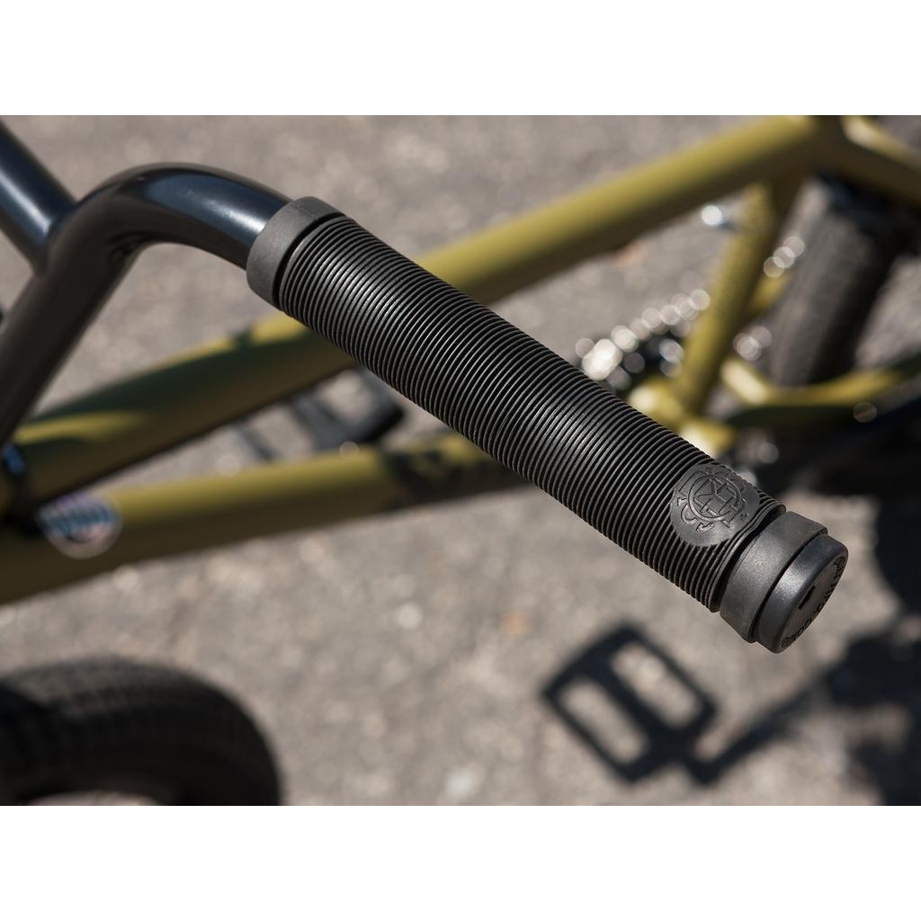 A close-up shows a textured black bicycle handlebar grip mounted on a Sunday Wave Length 20 Inch bike frame, styled like Gary Young's, over paved ground.