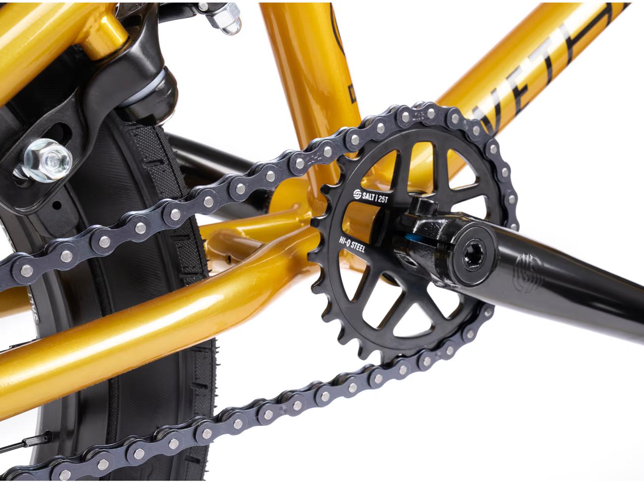 Close-up of a gold-painted bike frame featuring black Salt Rookie Tubular Chromoly Cranks, matching chainring and chain. A large tire tread is visible in the background, ideal for BMX enthusiasts or proud owners of a Wethepeople Seed 16 Inch Bike.