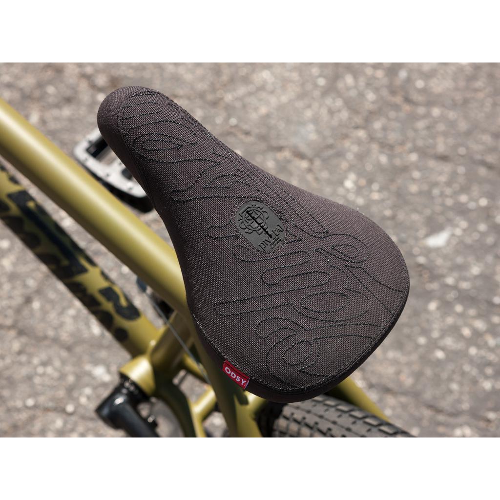 The bicycle seat has a textured black cover on an olive green frame with Gary Young Signature Chromoly parts from the Sunday Wave Length 20 Inch Bike, resting on a paved surface.