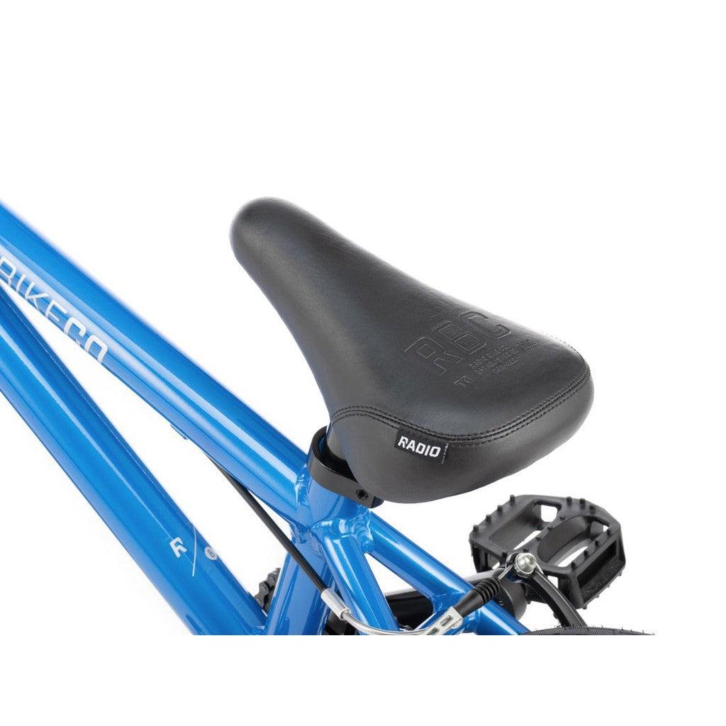 Close-up of a Radio Dice 14 Inch Bike's blue frame featuring a black saddle with "RADIO" and "RBC" branding, made from 6061-T6 alloy for a lightweight kids bike. A black pedal is visible in the background.