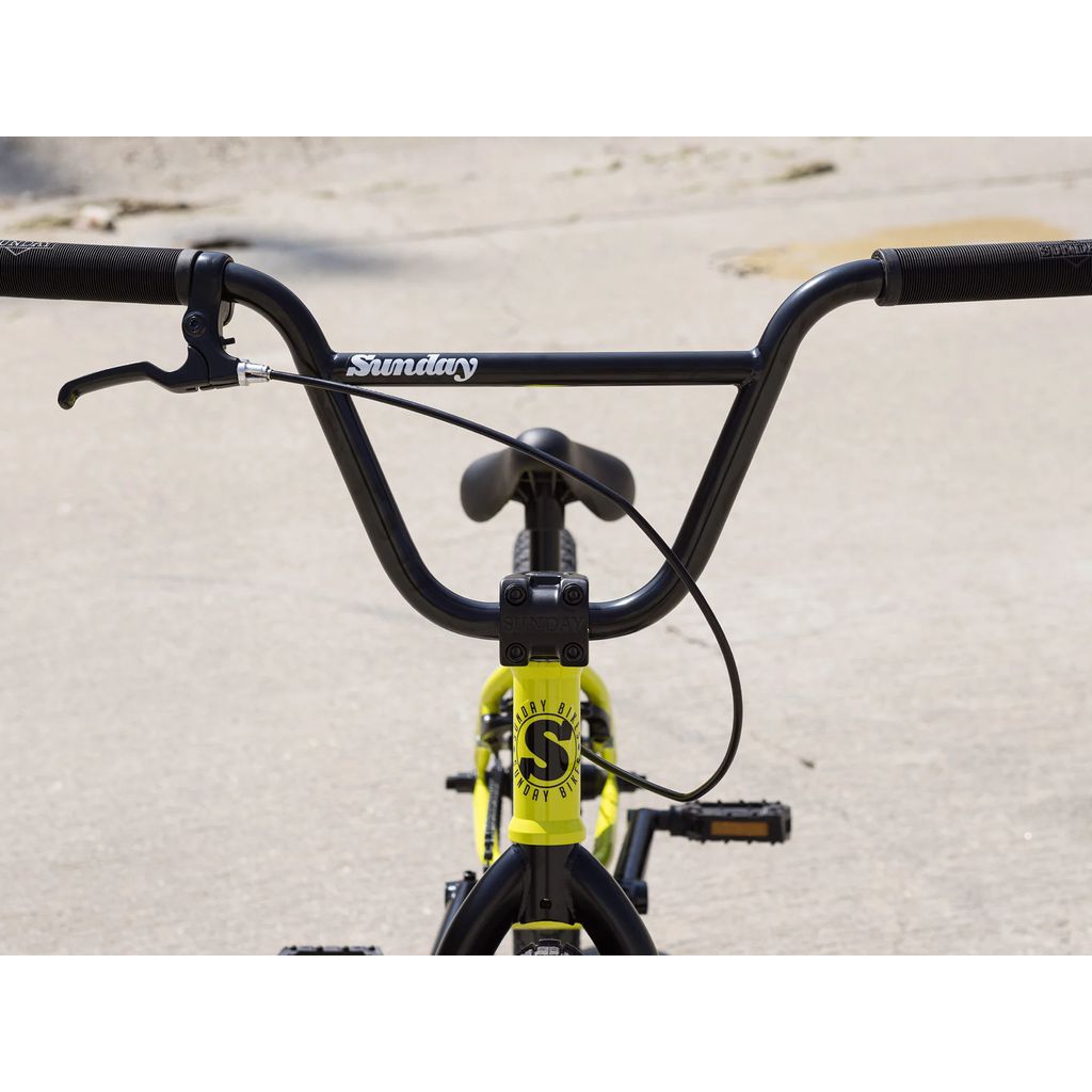 Close-up view of the handlebars of the Sunday Primer 18 Inch Bike (2025) with a black frame and partial yellow detailing, equipped with Odyssey Springfield brakes, standing on concrete.