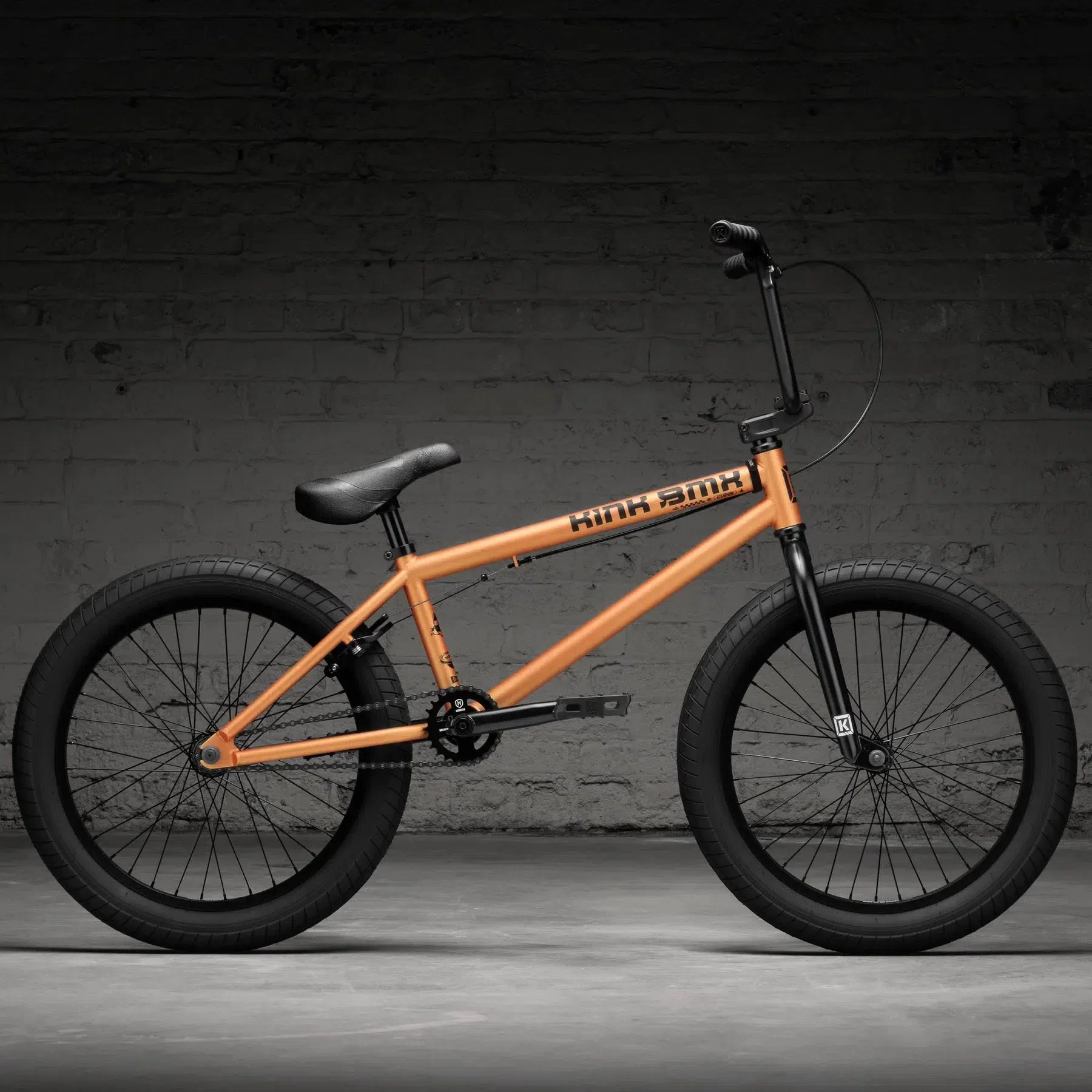 The Kink Curb 20 Inch BMX Bike (2026) is styled with an orange frame, black handlebars and seat, plus wide tires. It rests against a dimly lit brick wall background, making it perfect for entry-level riders aiming for an impressive start.