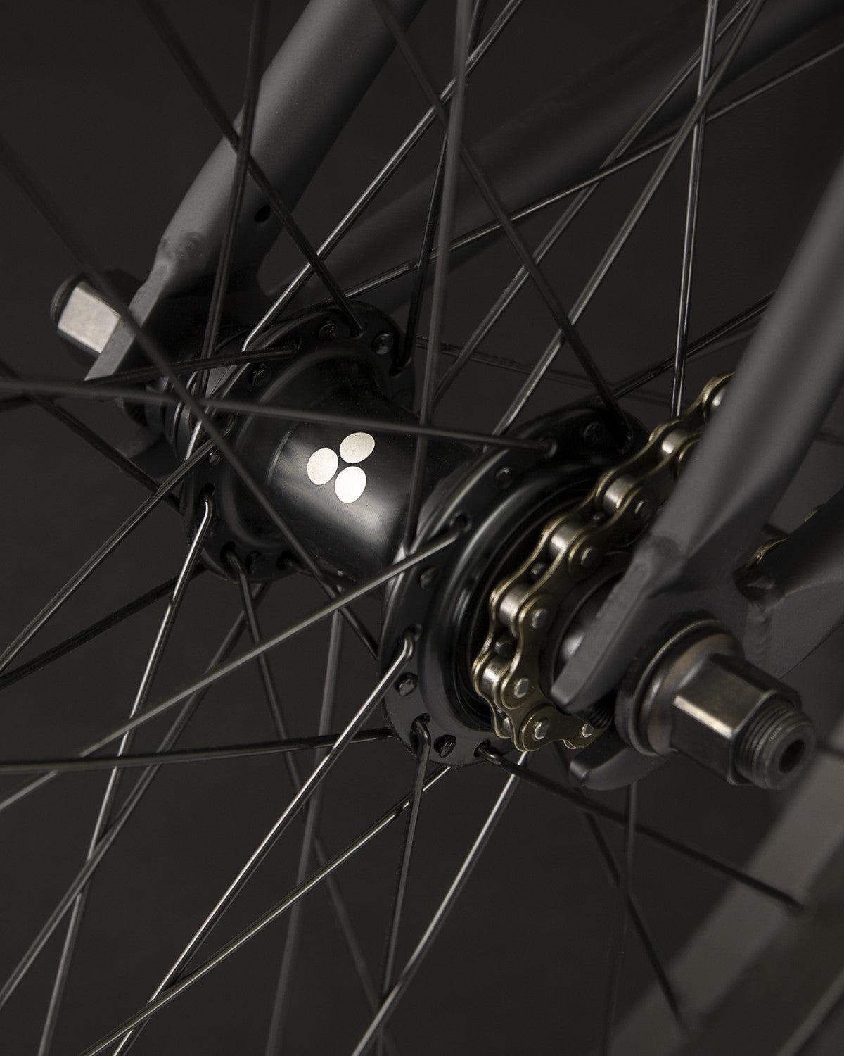 Close-up of the Fly Bikes Electron 20 Inch Bike wheel hub featuring the iconic three-dot logo, highlighting the durable Cr-Mo material, spokes, and chain mechanism in view.