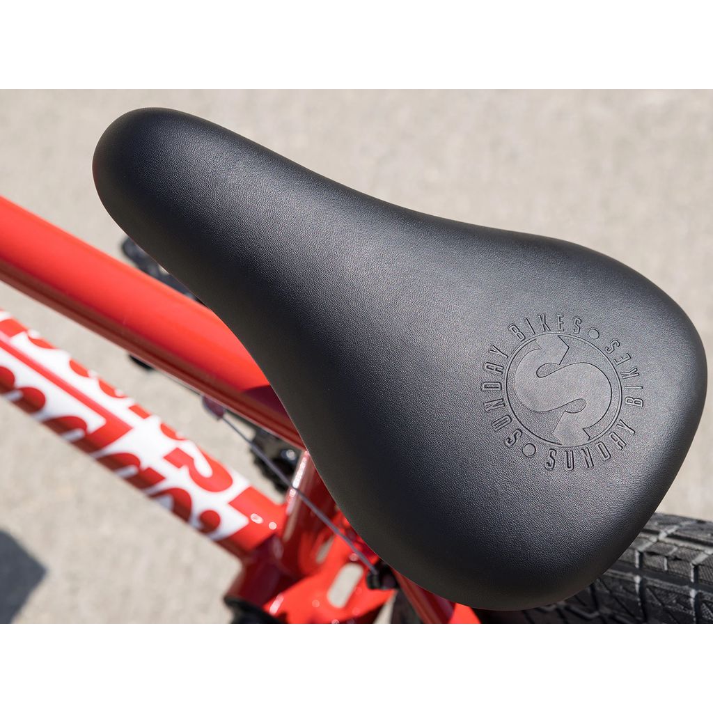 Close-up of a black bicycle seat mounted on the frame of a 2025 Sunday Primer 18 Inch Bike in red, adorned with the "Sunday Bikes" logo and featuring Odyssey Springfield brakes in the background.