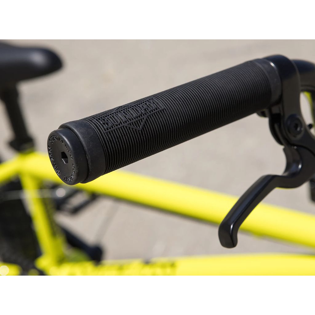 Close-up of a black handlebar grip and brake lever on a yellow BMX bike. The grip has a textured surface with branding, featuring Odyssey Springfield brakes on the Sunday Primer 18 Inch Bike (2025). The background is blurred.