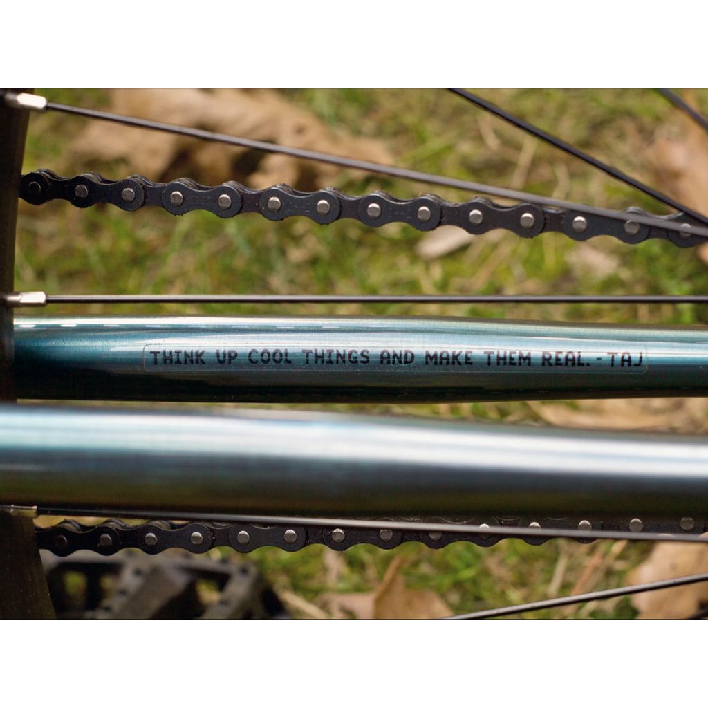 Close-up of a Fairdale Taj 27.5 Inch Bike (2022) frame, showcasing the text "Think up cool things and make them real. - Taj." Above the words, the single-speed drivetrain gleams, with grass subtly visible in the background.