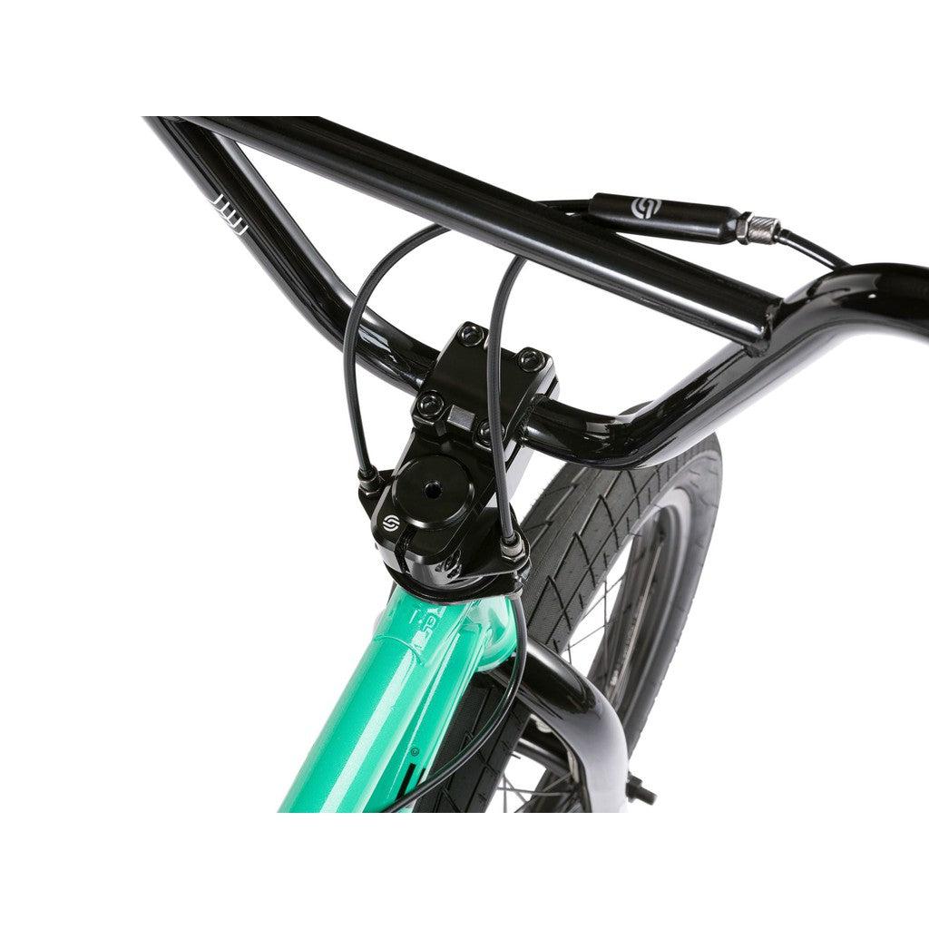 A close-up of the Wethepeople CRS FS 18 Inch BMX Bike showcases its black handlebars, brake cables, and turquoise frame near the front wheel.