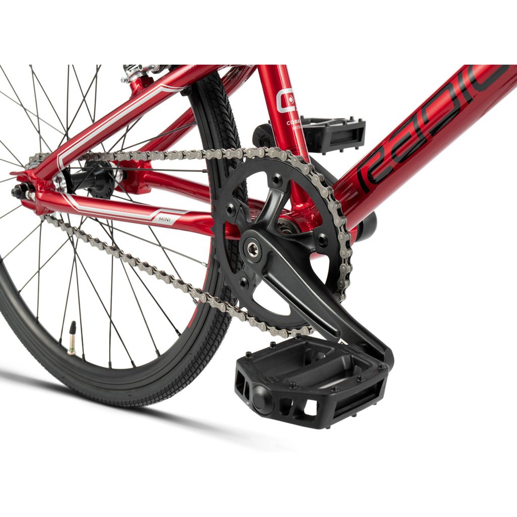 Close-up view of a red Radio Cobalt Mini Bike's chain and pedal system, showing the gears, chain, and a portion of the rear wheel. The bike has a sleek frame design reminiscent of BMX racing aesthetics, with clean mechanical components and precise engineering.