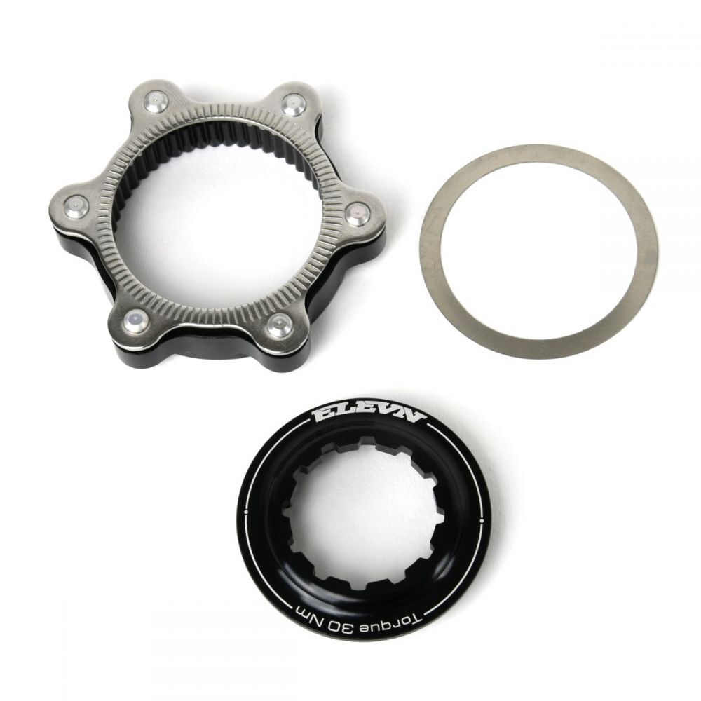 Three mechanical components are arranged on a white background: a star-shaped cog (gray and black), a flat circular metal ring (gray), and a cylindrical part labeled “ELEVEN Torque 30 NM” (black) from the Elevn Centerlock ISO Adapter Kit.