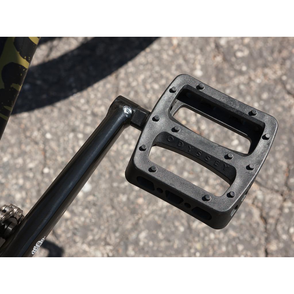 Close-up of a black bicycle pedal with "Odyssey" branding, attached to a crank arm of the Sunday Wave Length 20 Inch (Gary Young) Bike, resting on an asphalt surface.