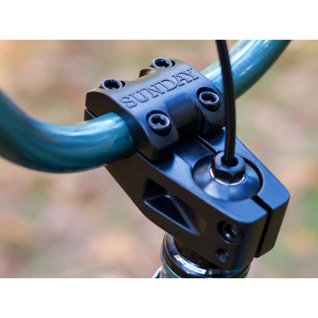 Close-up of a black bicycle handlebar stem engraved with "Sunday" on the sleek Fairdale Taj 27.5 Inch Bike (2022). The handlebars are teal, while the background is artfully blurred, showcasing its elegant chromoly frame and single-speed drivetrain.