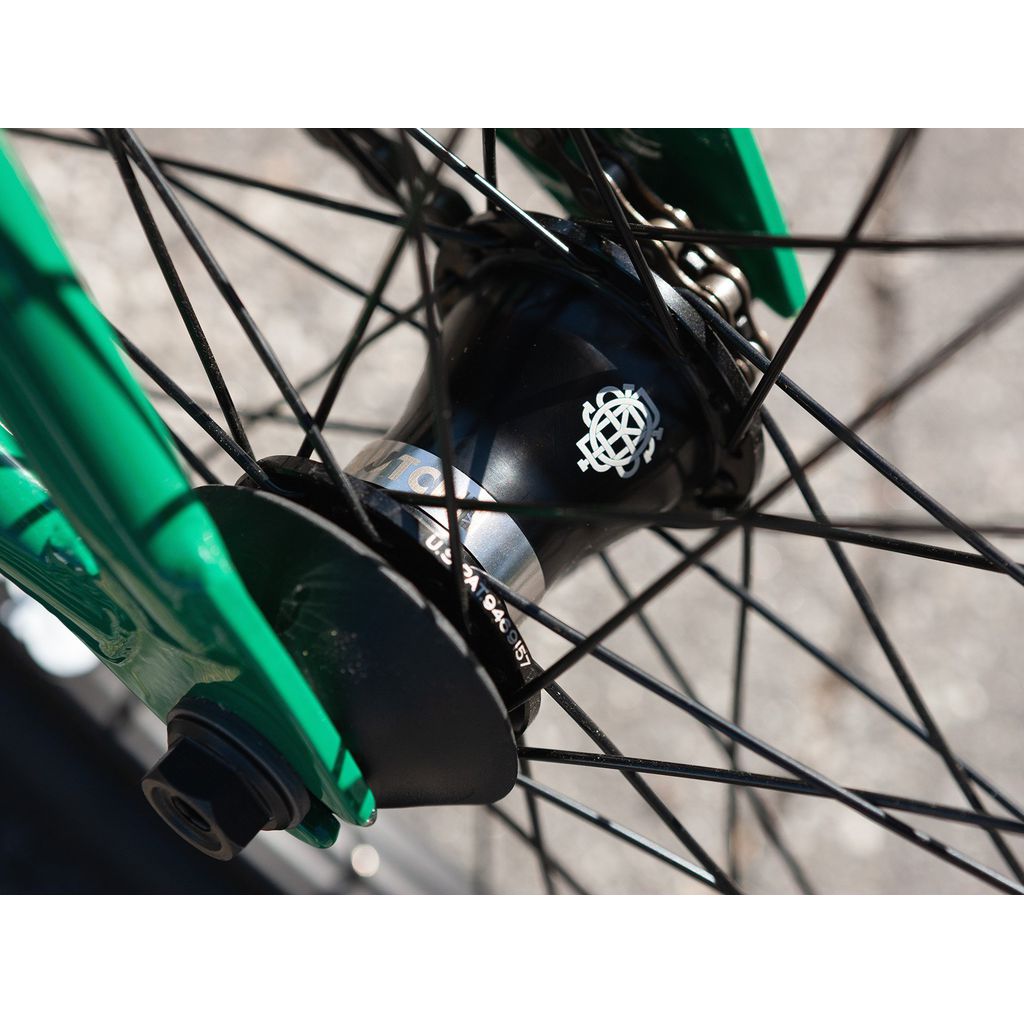 Close-up view of the hub area of the Sunday Forecaster 20 Inch Bike (2022) / Alec Siemon, showcasing the black Odyssey Clutch v2 Freecoaster hub shell with a printed logo, black spokes, and a portion of a green fork. The background is a blurry gray surface.