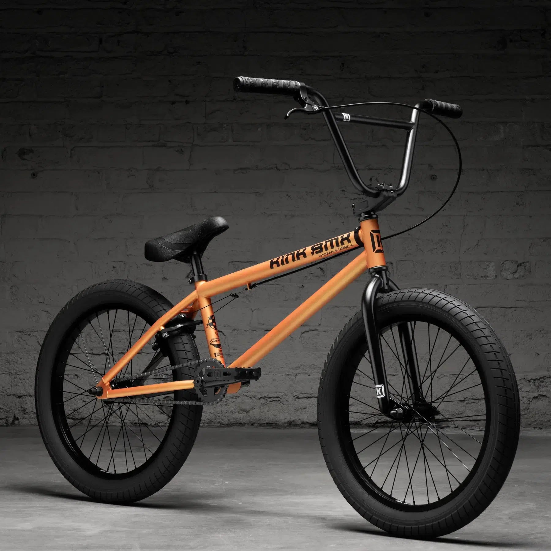 The Kink Curb 20 Inch Bike (2026) features an orange frame, black handlebars, and wide tires. It's set against a concrete floor and brick wall backdrop, making it ideal for entry-level riders.