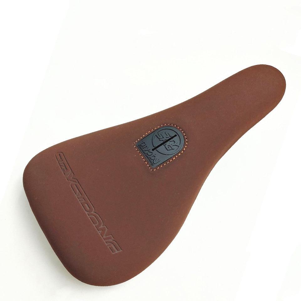 Brown bicycle seat with a smooth microfibre surface, showcasing a small black emblem and the word "SAVEDOUBLE" embossed on the side. The Stay Strong Cut Off Slim Pivotal Seat design ensures durability throughout your ride.