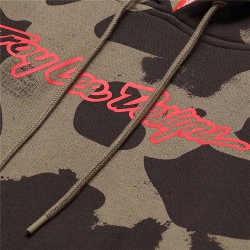 Close-up of a camouflage TLD 25.1 Signature Pull Over Hoodie featuring "Lee Jeans" in red cursive on the chest. Brown drawstrings enhance its rugged charm, making it a wardrobe staple.