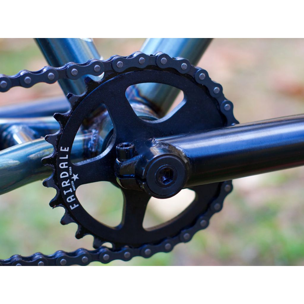 Close-up of a bicycle crankset featuring a Fairdale chainring. The black chain and crank are integral components of the single-speed drivetrain, linked to the teal chromoly frame of the Fairdale Taj 27.5 Inch Bike (2022).