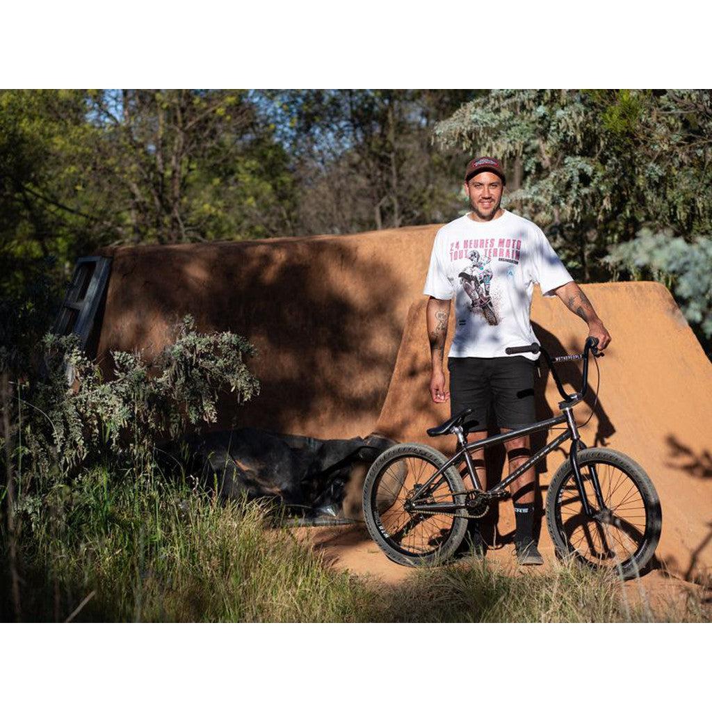 A person stands outdoors beside a Wethepeople Chaos Machine 22 Frame bicycle in front of a dirt ramp and trees, wearing a white graphic t-shirt and black shorts.