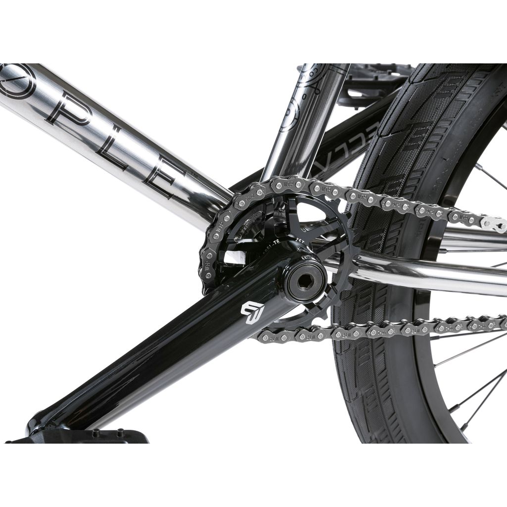 Close-up of a BMX bike's drive train, featuring a silver chain, black crank arm, and large gear teeth on a chromoly frame. Part of the rear wheel and tire from the Wethepeople Envy 20 Inch BMX Bike is also visible.