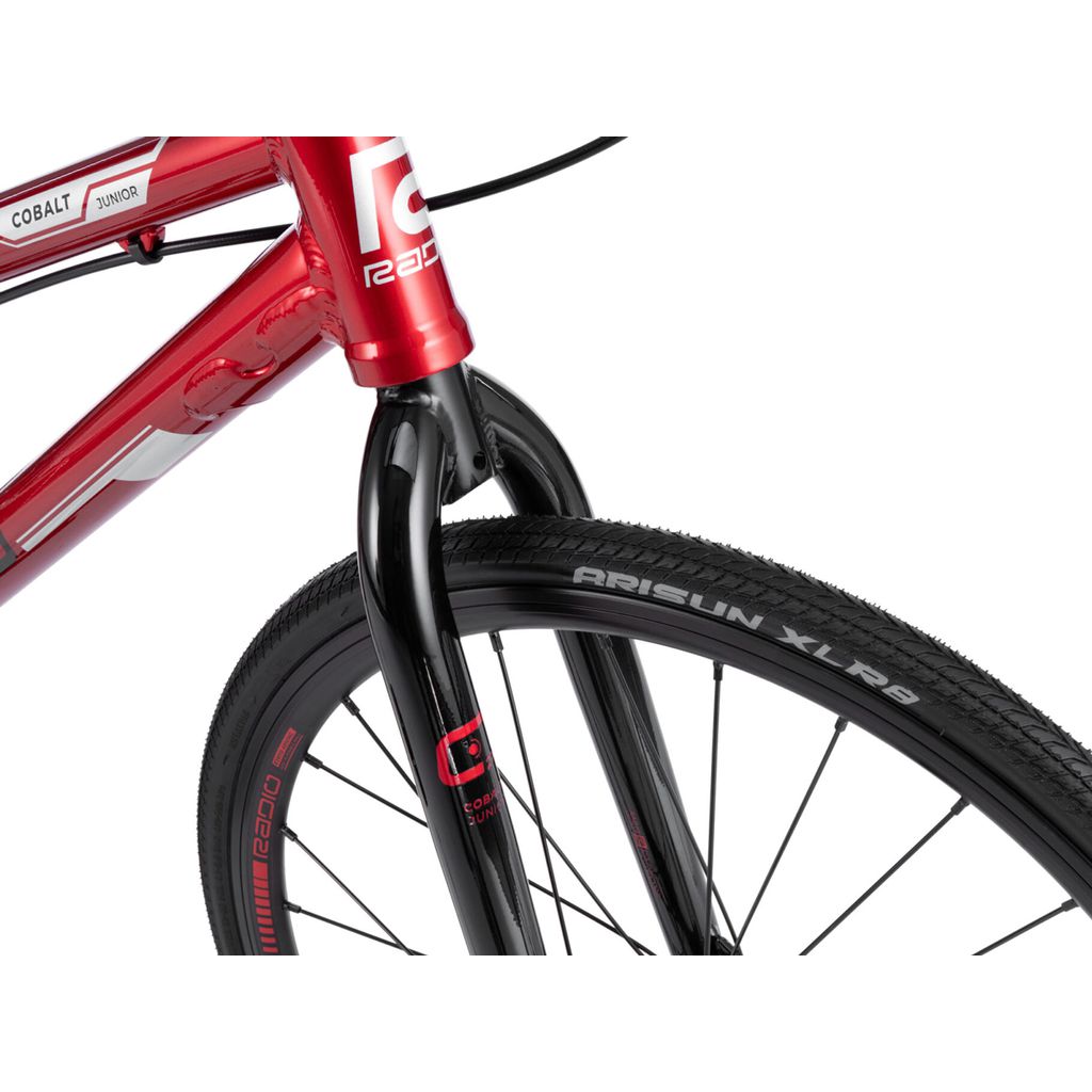 Close-up view of the front section of a red BMX race bike, showing the fork, tire, and part of the frame. The tire is branded "ARISUN XLR8" and the frame has a "Radio Cobalt Junior Bike" label from Cobalt bikes.