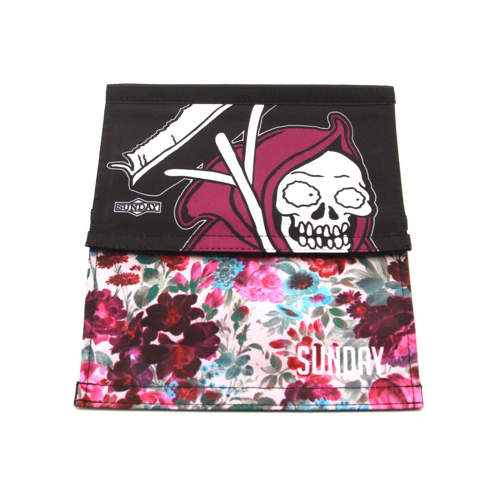 Two fabric face masks, one showcasing a skull and scythe design against a black backdrop and the other adorned with a floral pattern, are displayed unfolded. Featuring the distinct 2-in-1 style of the Sunday Wallflower V2 Reversible Handlebar Pad, both masks prominently feature the word "SUNDAY.