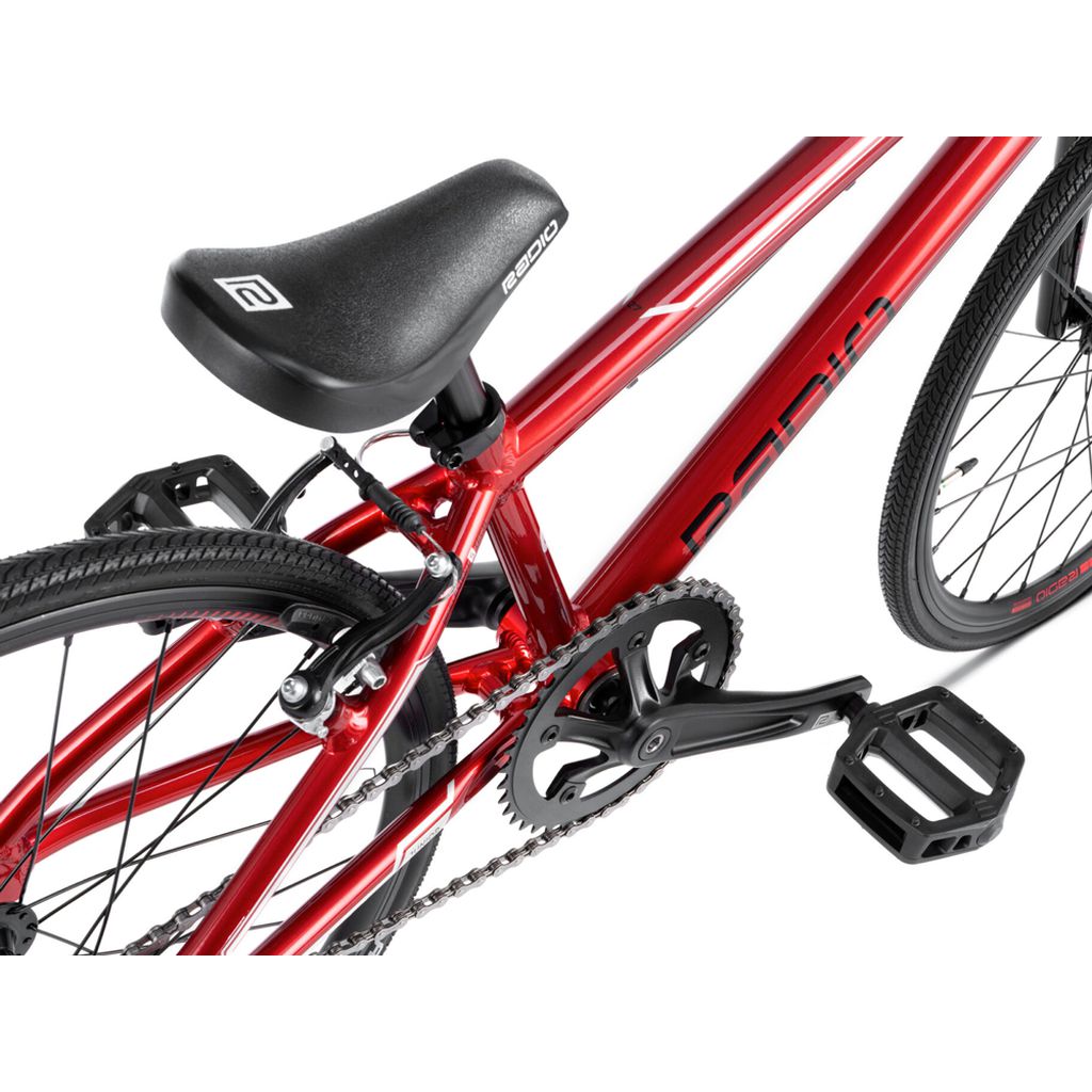 Red bicycle with a close-up view of its black saddle, pedal, and drivetrain components. The bike, a Radio Cobalt Junior Bike, has a sleek frame design and detailed features reflecting top-notch craftsmanship.