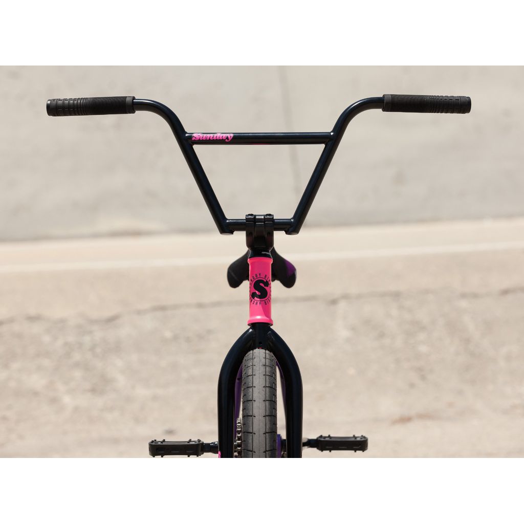 The front view of the Sunday Street Sweeper 20 Inch Bike showcases black handlebars and a pink stem against a concrete backdrop. Its robust chromoly frame complements the sleek design, while the Odyssey Clutch freecoaster ensures an unmatched riding experience.