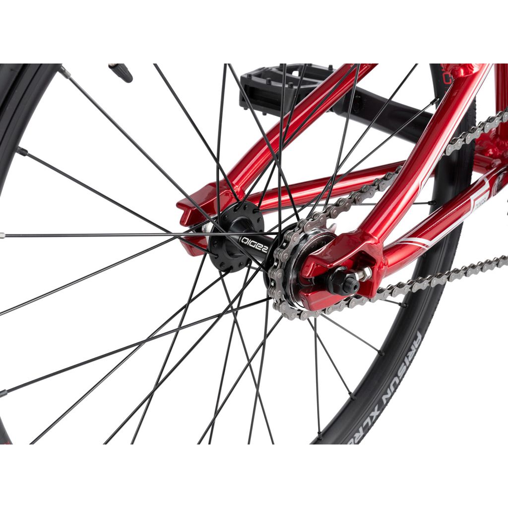 Close-up of a Radio Cobalt Junior Bike's rear wheel and gears with black spokes, a red metal frame, and a tire on a ruler for calibration.