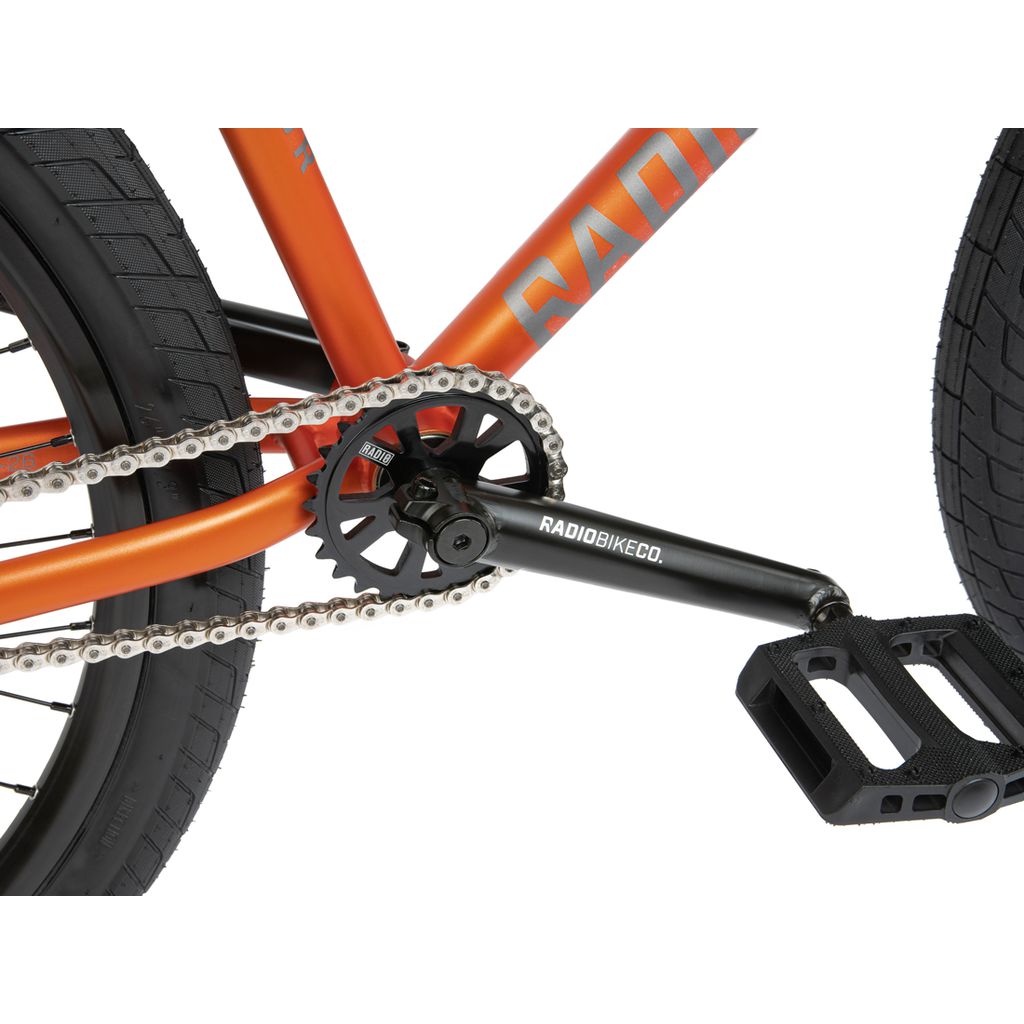 Close-up of an orange BMX bike's drivetrain, including the chain, chainring, crank arm, and pedal equipped with sealed bearings. The text "Radio Bike Co." is visible on the crank arm of the Radio 26 Inch Ceptor Bike.