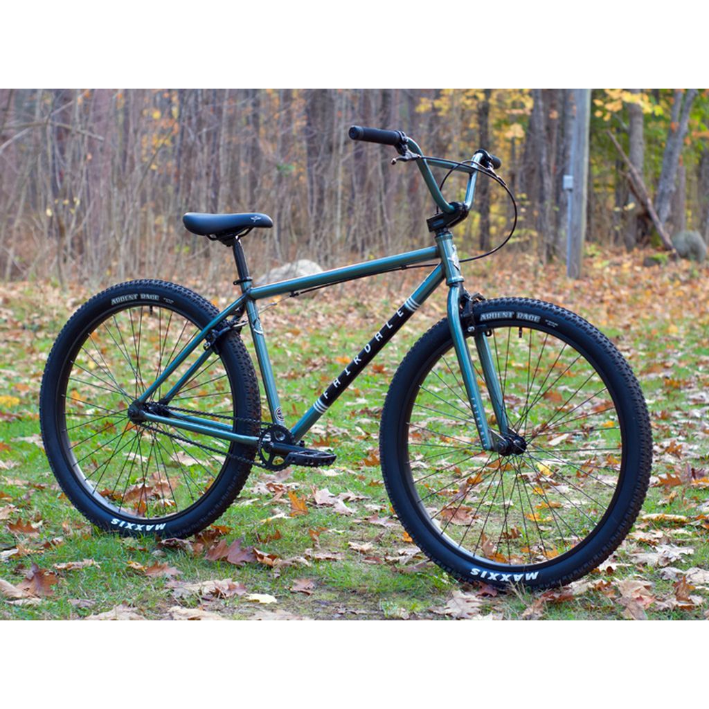 A Fairdale Taj 27.5 Inch Bike (2022) in metallic green with black tires and a straight handlebar rests on the grass, showcasing its chromoly frame and single-speed drivetrain, set against a backdrop of autumn trees and scattered leaves.