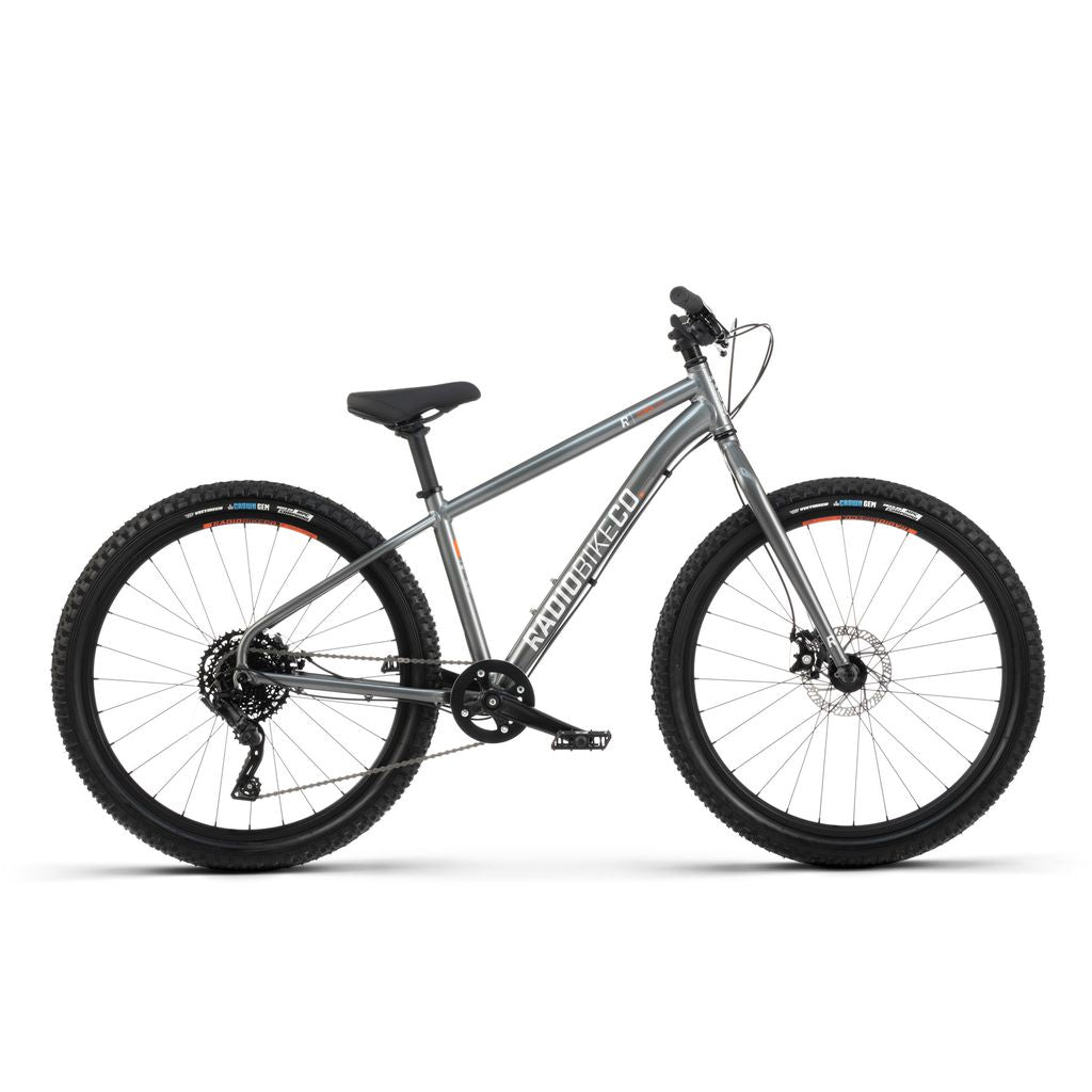 The Radio 26 Inch Zuma Bike, a silver-grey Kids MTB bike featuring a lightweight alloy frame, front suspension fork, knobby tires, and a black seat, is displayed against a plain white background.