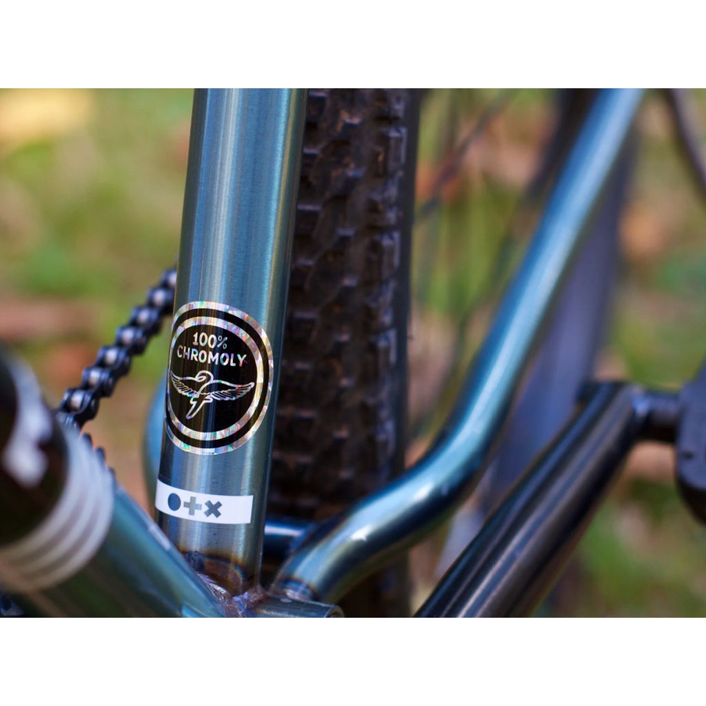 Close-up of the Fairdale Taj 27.5 Inch Bike (2022) reveals a "100% Chromoly" sticker on its robust chromoly frame, right next to the tire, showcasing its sturdy single-speed drivetrain.