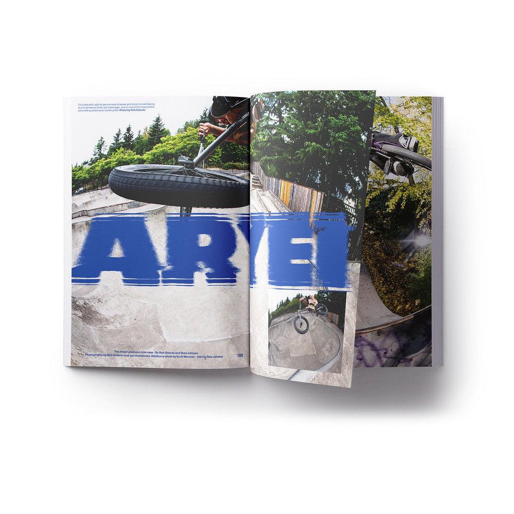 In the DIG Book 2024 - Photo Annual, a BMX rider performs stunts in a skate park, framed by trees and a fence. The word "AVEL" is prominently displayed in large blue text across the pages.