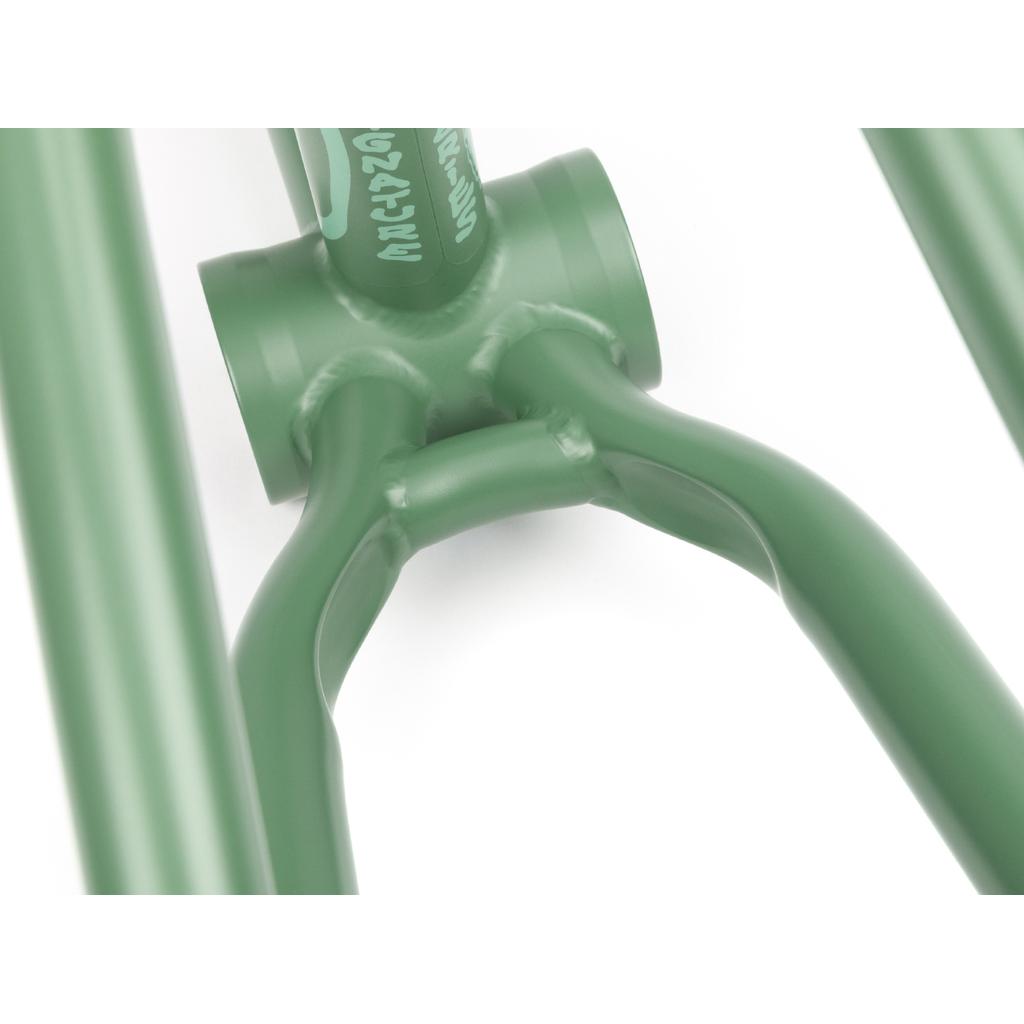 Close-up of a green bicycle frame joint highlighting welds and curved metal tubes. The modern street design is evident, featuring partial white text on what must be a BSD Jonesin Frame (2021).