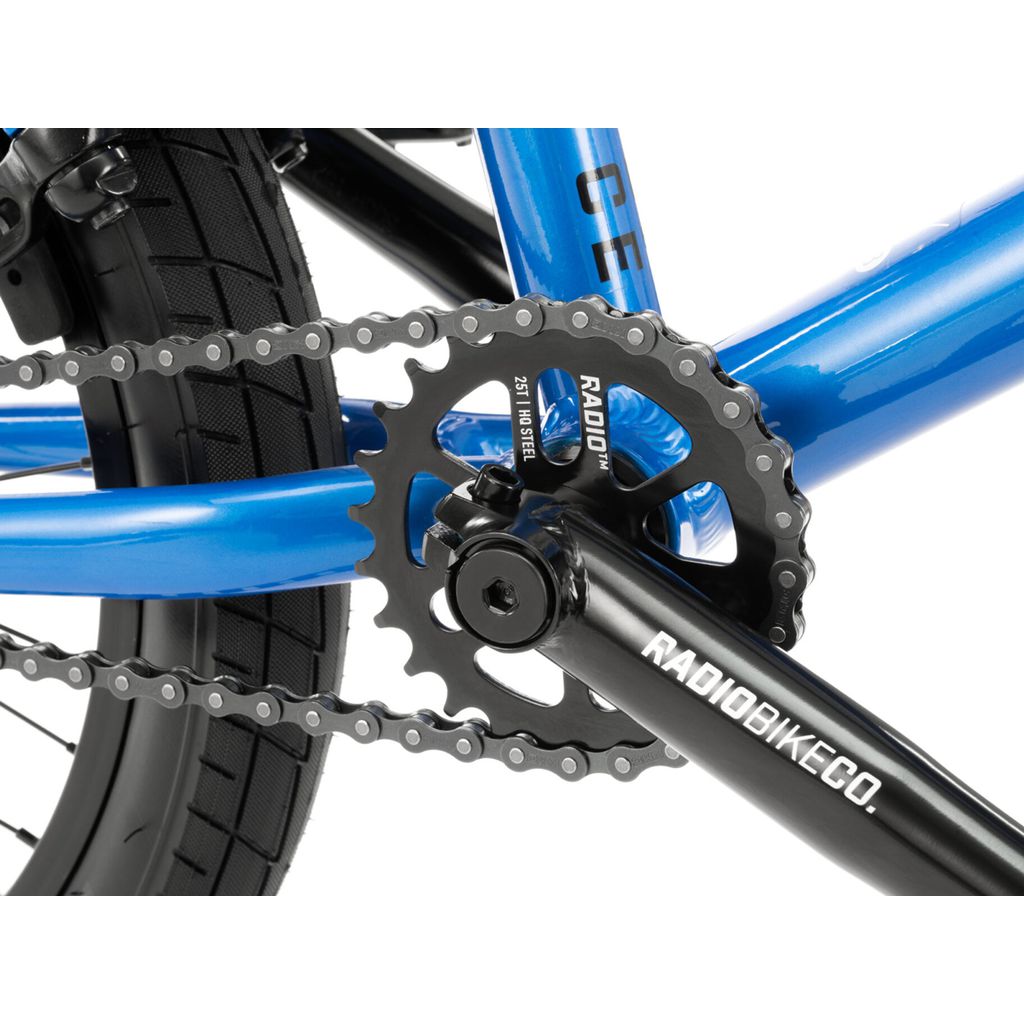 Close-up of a blue Radio Dice 18 Inch Bike's drivetrain showcasing the chain, black chainring labeled "RADIOBIKECO," and part of the tire and frame. The bike is equipped with Radio Rookie Three Piece Cranks for added durability.