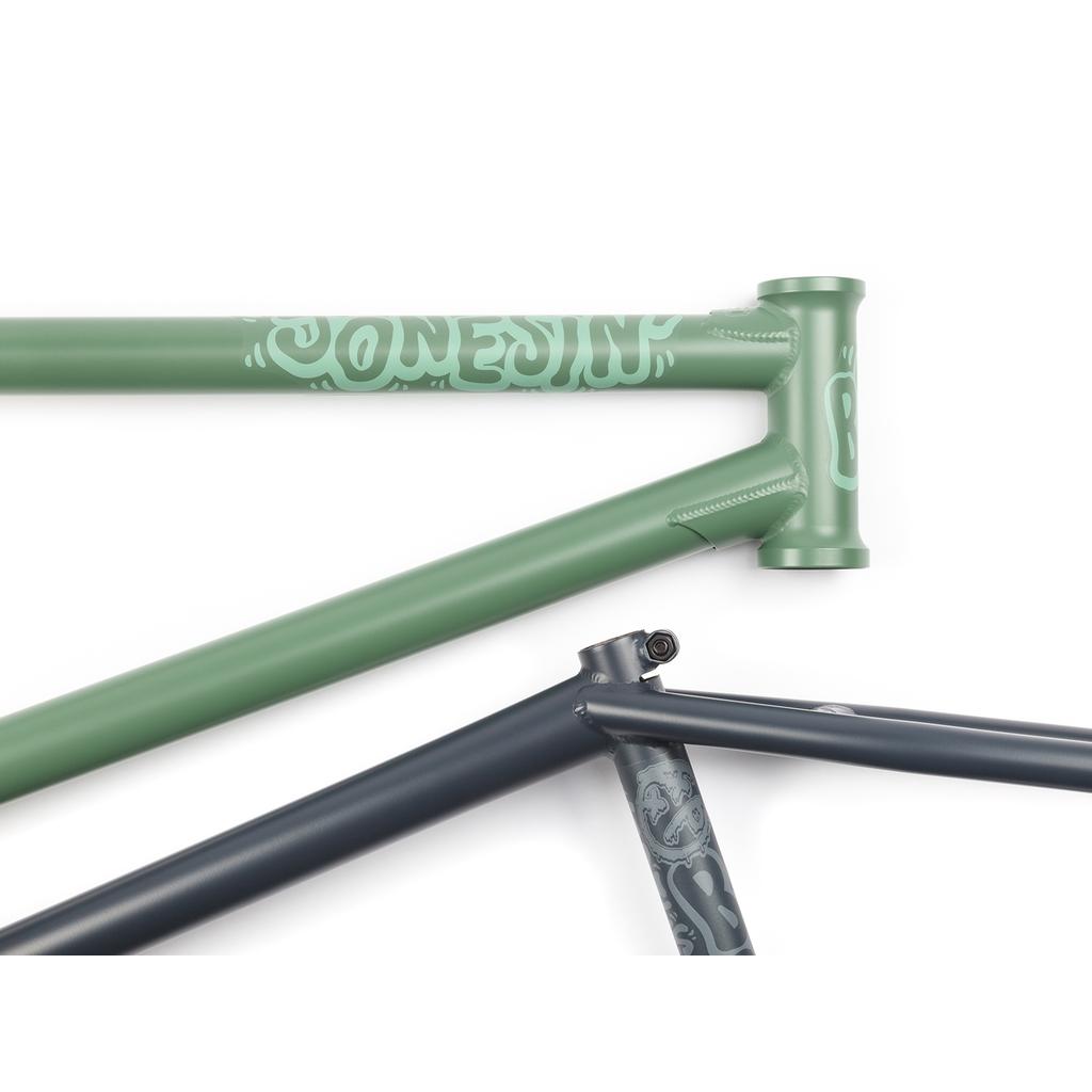 Close-up of two bicycle frames: one is a matte green BSD Jonesin Frame (2021) with white lettering honoring Sam Jones, and the other is dark gray with faint designs.