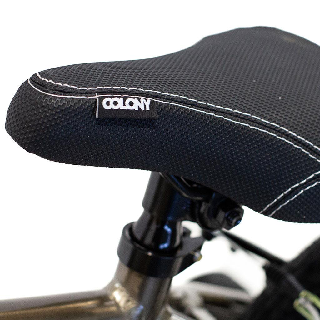 Close-up of a black and grey textured bicycle seat with white stitching from Colony, mounted on a sleek BMX bike. The lightweight alloy frame of the Colony Horizon 12 Inch Bike is partially visible.