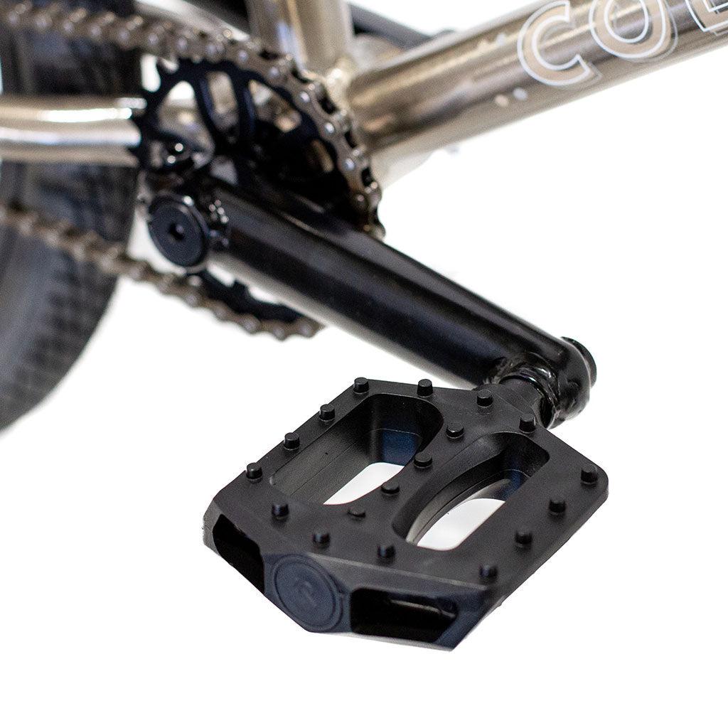 Close-up of a BMX bike pedal and crank arm attached to the bottom bracket of the Colony Horizon 20 Inch Freestyle Bike. The black pedal features metal pins for grip, ensuring firm footing for any freestyle Olympic athlete. The bike's chain and part of the frame are also visible.