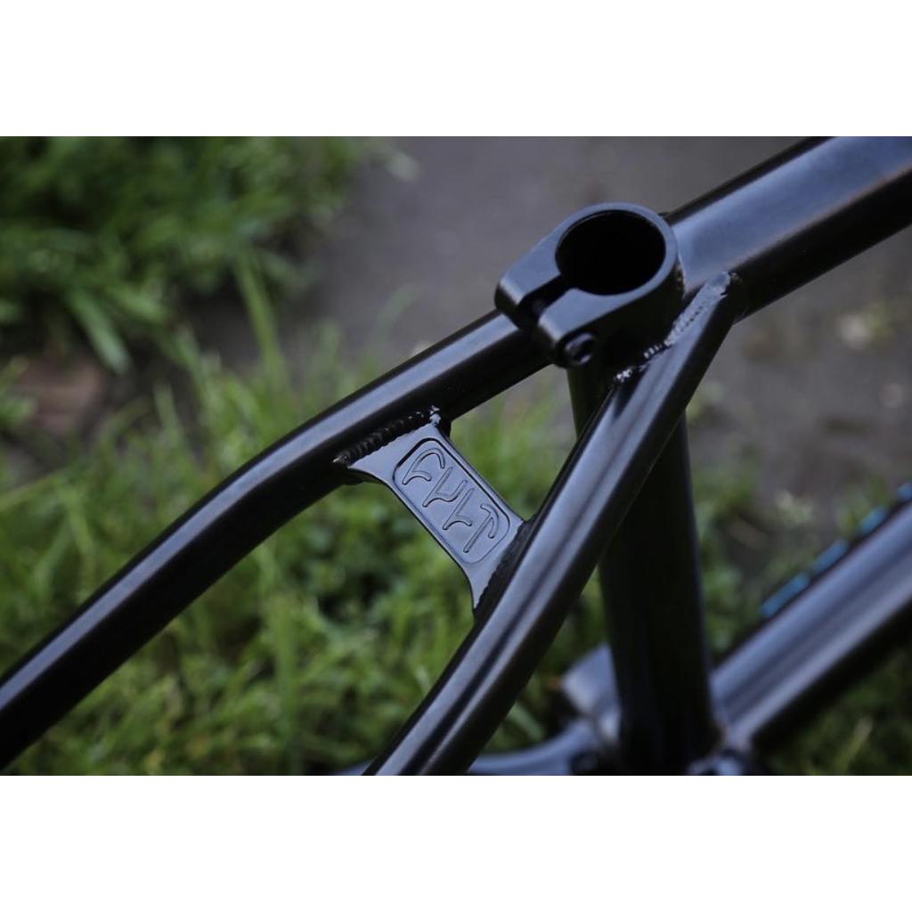 Close-up of the Cult 2 Short IC Frame, showing "FYP" engraved on an investment cast metal support bar. The background of grass and pavement highlights the craftsmanship of this cult classic tubing.