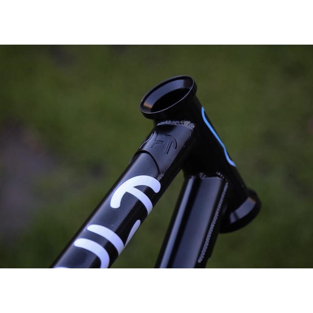 A black Cult 2 Short IC Frame with white markings is featured close-up, highlighting its classic tubing on the head tube and part of the top tube, set against a blurred green background.