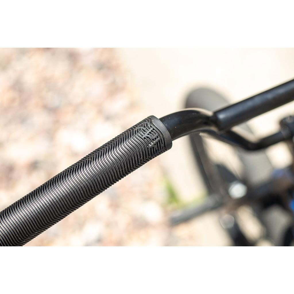 Close-up of textured black Cult Ricany Grips on a bicycle handlebar, set against a blurred gravel background.